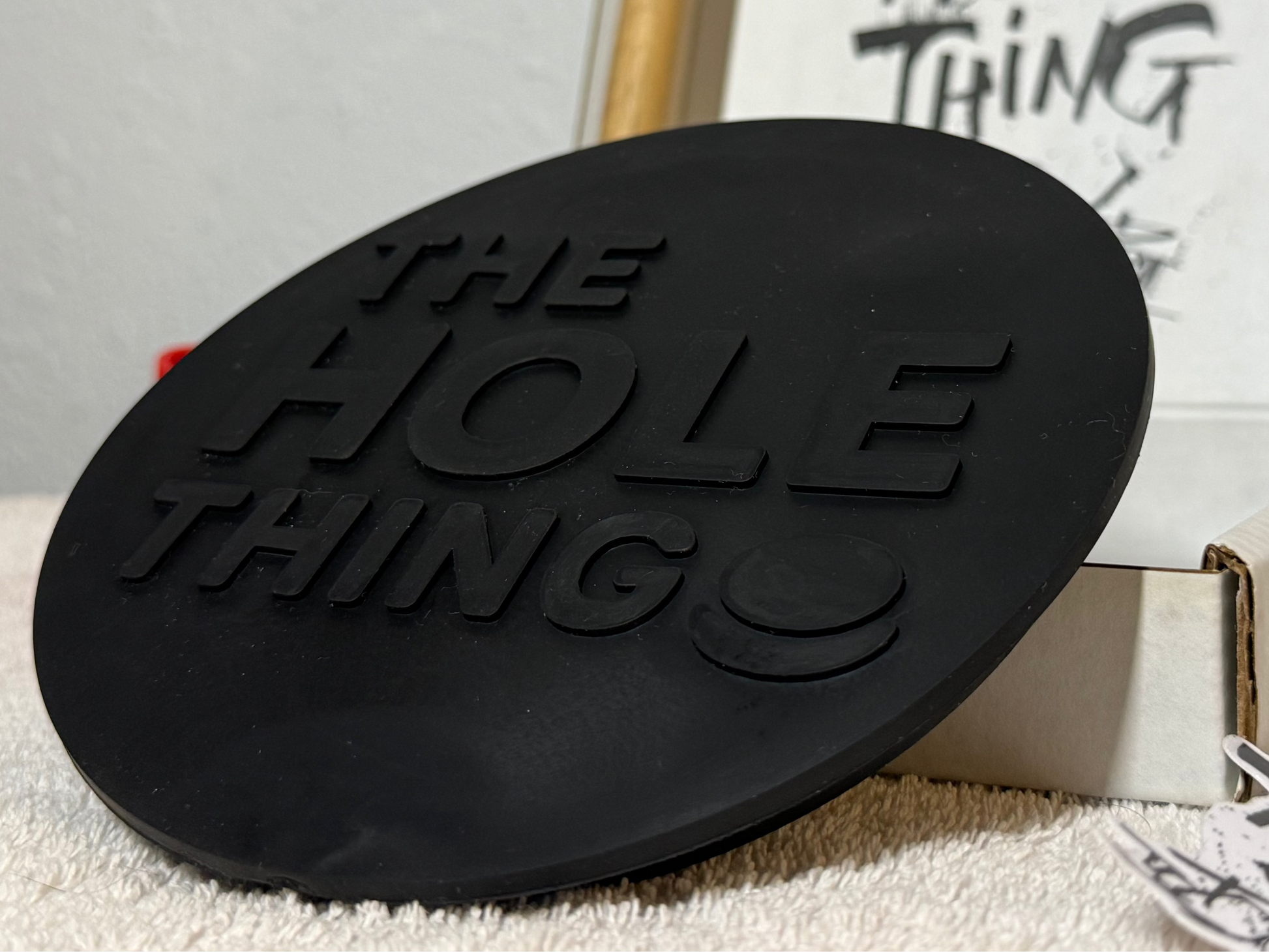 A round black rubber or silicone coaster with embossed text reading 'THE HOLE THING' sits in front of a white box containing a matching printed card or insert. The scene is photographed indoors on a light textured surface against a plain wall.